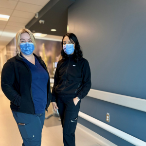 Two female nurses wearing masks and standing together in hallway.