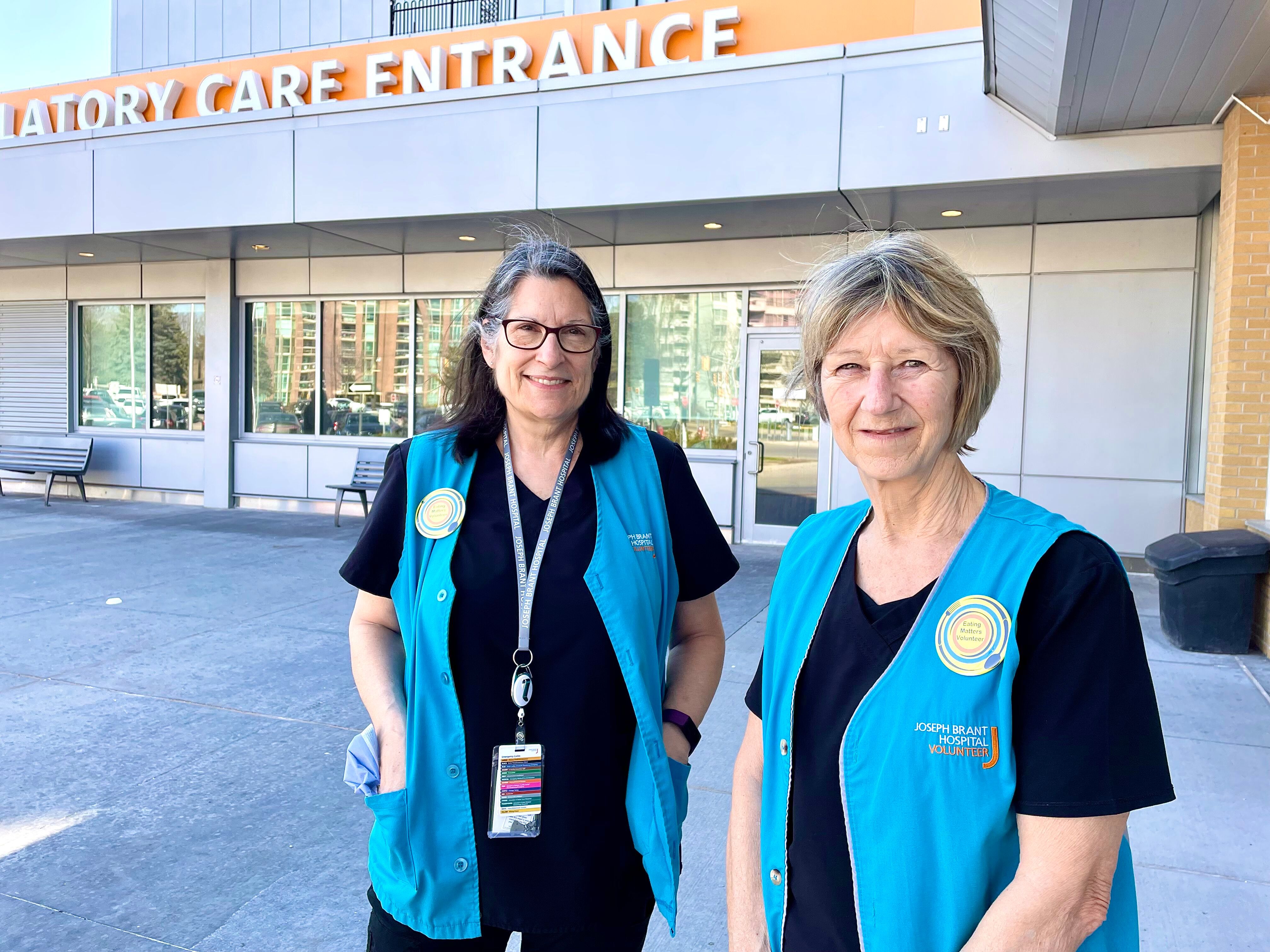 Two female volunteers wearing blue vests standing together outside of the hospital.