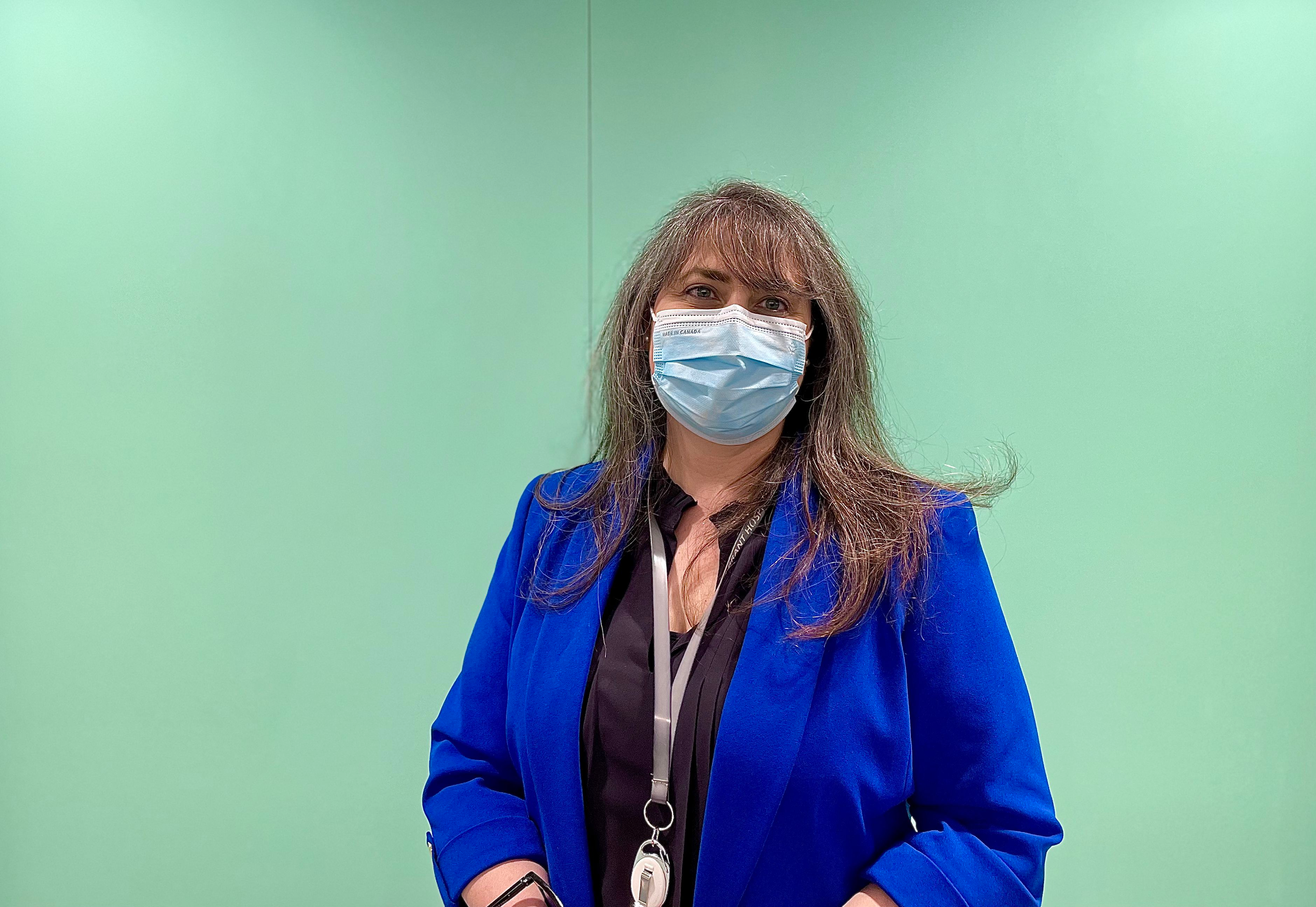 Anastasia Taylor standing in front of light blue wall wearing a dark blue blazer, black shirt and mask over her face.